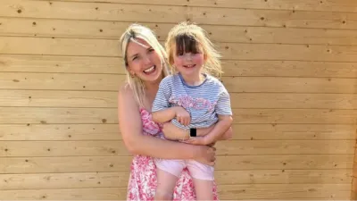 A mother and her daughter pose outside against a wooden panel wall.