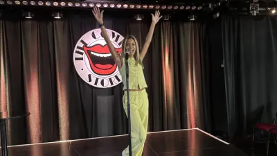 A comedian stands on stage at the Comedy Store raising her arms.