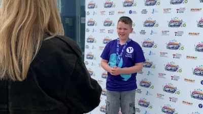 A boy in a Young Epilepsy t-shirt stands in front of a CarFest step and repeat.