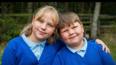 A brother and sister sit on a bench in their school uniforms.