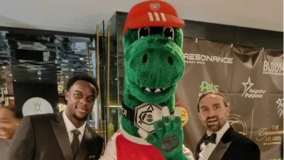 Two young men in suits stand with an Emirate dinosaur mascot at the Empowerment Awards.