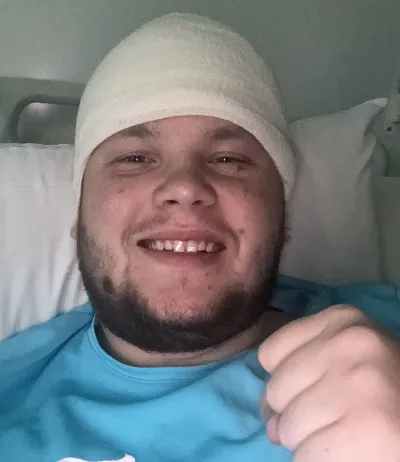 A young man in a head bandage fist bumps camera from a hospital bed.