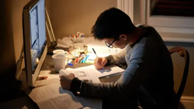 A young boy studies at a computer with a cluttered desktop at night time.