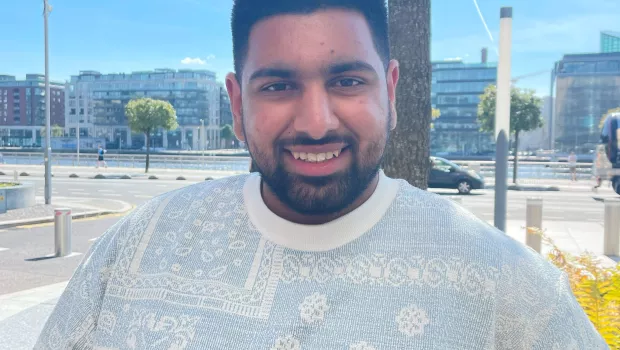 A bearded young man in a white t-shirt smiles into the camera on a summery day.