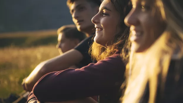 A group of teenagers sit smiling in a row outside in the low light of sunset.