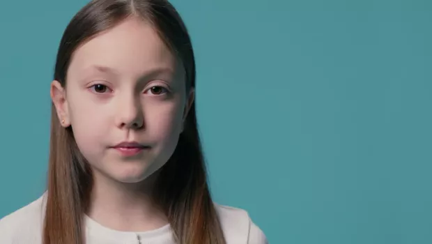 A young Caucasian girl stands against a blue backdrop wearing a white t-shirt