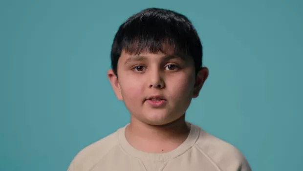 A young Asian boy stands against a blue backdrop wearing a white t-shirt