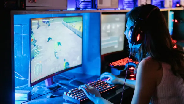 A young woman smiles as she plays video games in a computer room, wearing a headset.
