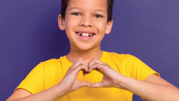 Young boy smiling in a yellow t-shirt holding a hand heart against a purple backdrop.