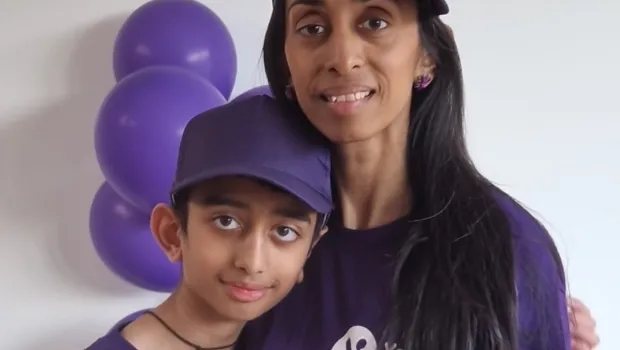 A mother and son in Young Epilepsy clothes smile before purple balloon, wearing Young Epilepsy merchandise.