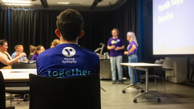 A young man in a Young Epilepsy shirt, pictured from behind, watches a lecture from his seat.