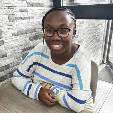 Photograph of young woman sat at a table, arms cross, smiling at the camera 