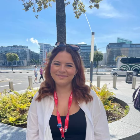 Photograph of young woman smiling at the camera, with a backdrop of  blue sky and office buildings. and .  