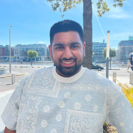 A bearded young man in a white t-shirt smiles into the camera on a summery day.