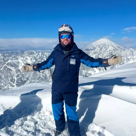 Miles, a young man dressed in snowboarding attire, smiles and opens his arms to camera atop a snowy mountain.