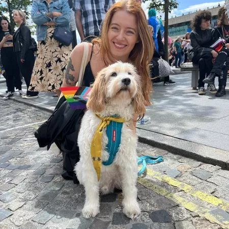 A young woman holding pride flags smiles next to a dog on a sunny day.