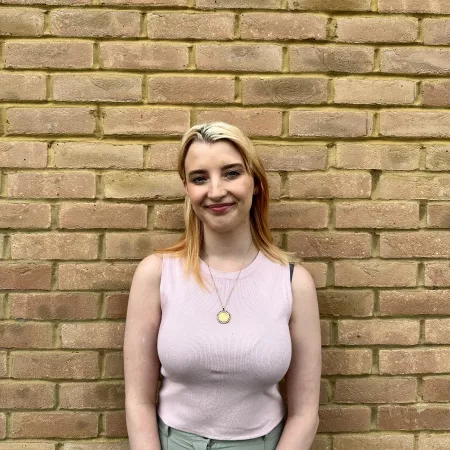 Bethany, a young blonde woman, stands smiling against a brick wall.