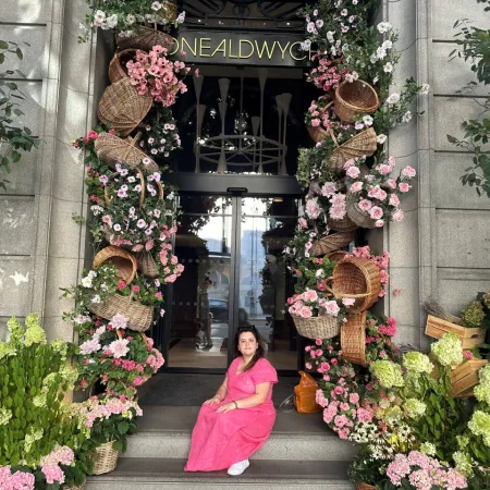 A young woman sits on the step of a doorway, surrounded by baskets of pink flowers.