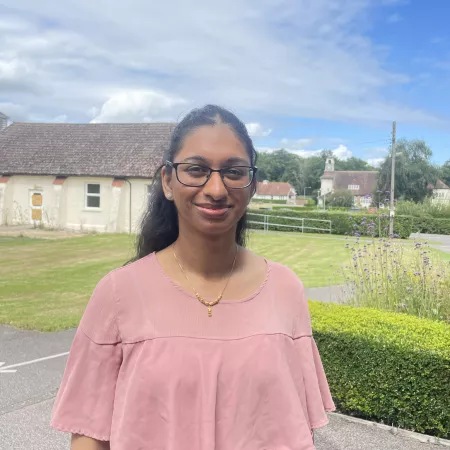 A young woman, Tee, smiles in a rural area, wearing glasses and a peach blouse.