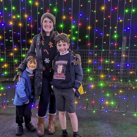 A mother and her two sons smile at Christmas time in front of a wall of fairy lights.