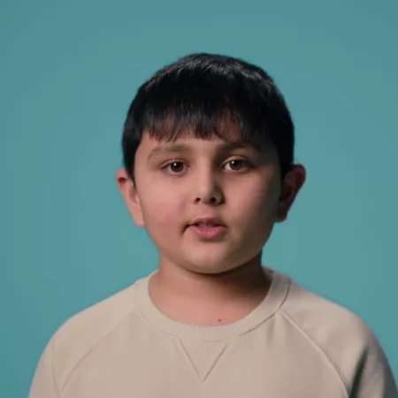 A young Asian boy stands against a blue backdrop wearing a white t-shirt