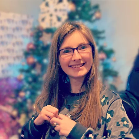 A young woman in glasses and pyjamas smiles before a Christmas tree.