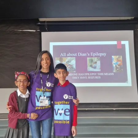 A mother stands with her two children in Young Epilepsy t-shirts in front of a screen.