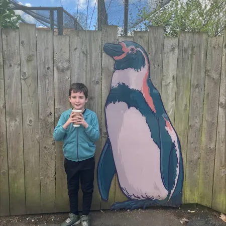 A young boy, Tate, stands beside a fence with a painting of a penguin.