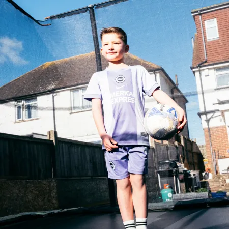 A young boy stands on a trampoline holding a football. 