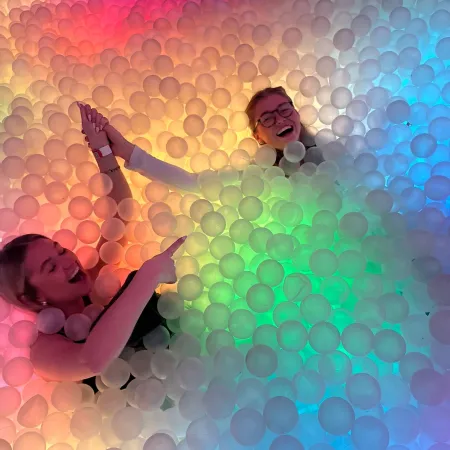 Two women lie down in a rainbow-themed ball pit.