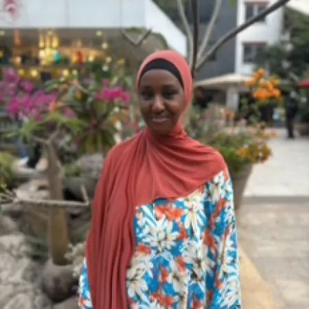 A young woman in a headscarf smiles in a leafy street.