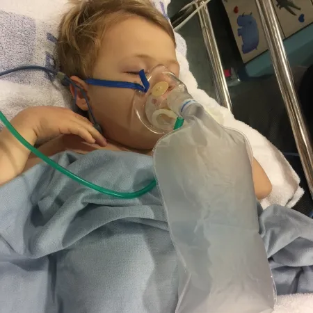 A boy lies in a hospital bed with a breathing mask.