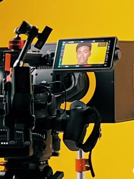 Photograph of teenager on a film set.  The back drop is yellow and he is sat on a stool talking to the camera. 