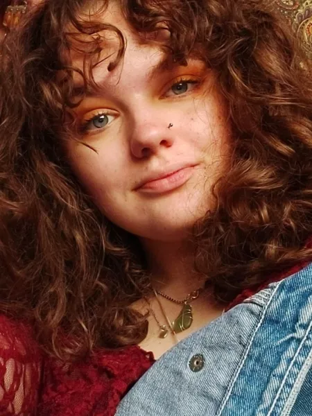 Daisy, a young woman with curly hair wearing denims, poses for a selfie.