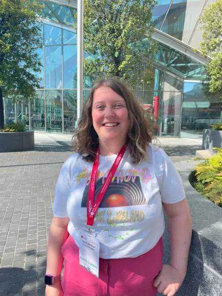 A young woman named Annie smiles to camera before a glass building on a sunny day.