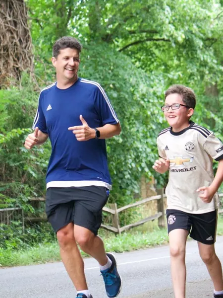 A young boy in glasses runs alongside his father on a tree-lined street, both smiling.