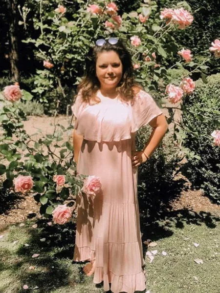 A young woman in a peach dress and sunglasses stands in a rose garden.