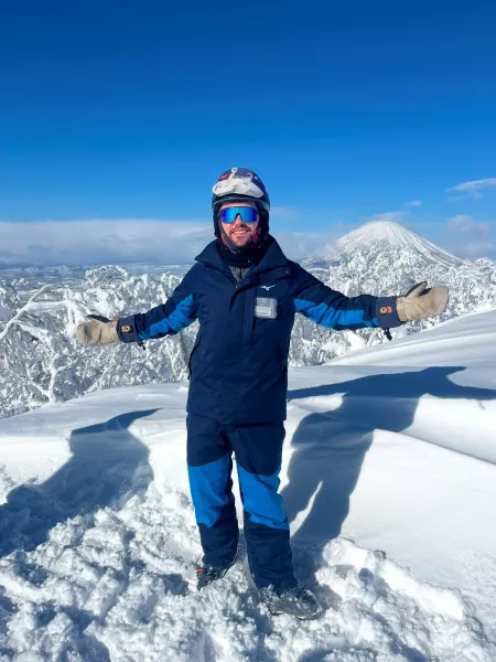 Miles, a young man dressed in snowboarding attire, smiles and opens his arms to camera atop a snowy mountain.