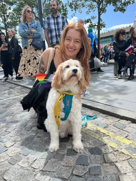 A young woman holding pride flags smiles next to a dog on a sunny day.