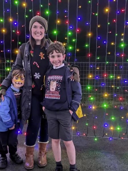 A mother and her two sons smile at Christmas time in front of a wall of fairy lights.