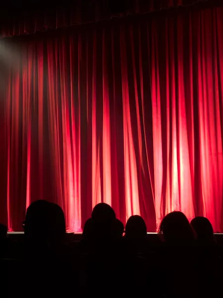 A dark crowd looks at closed red theatre curtains.