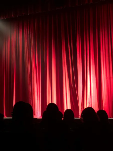 The silhouette of an audience in shadow look to a red stage curtain.
