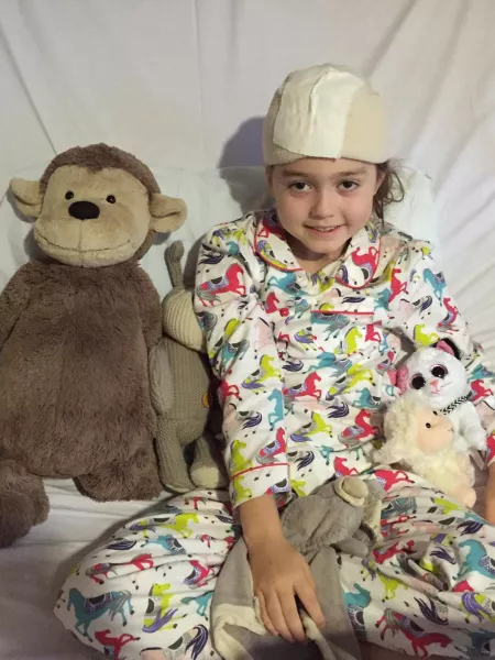 A young girl with a surgery bandage on her head sits with her stuffed teddies.