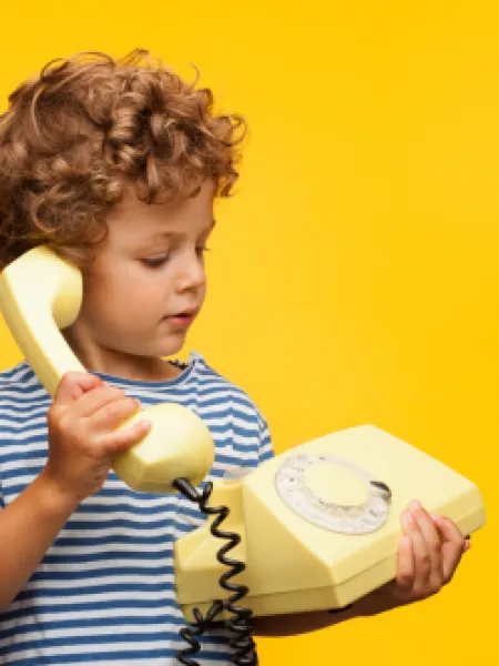 A young boy against a yellow backdrop holds a traditional phone in his hand and headset to his ear.