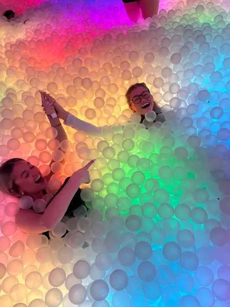 Two women lie down in a rainbow-themed ball pit.