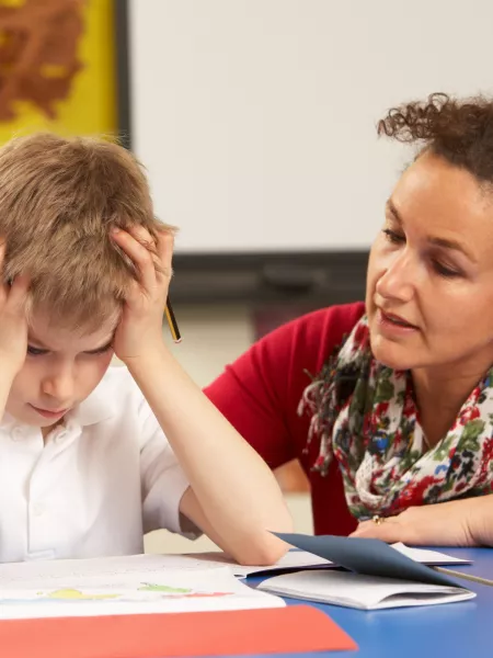 An educator speaks gently to a young student, who is holding his hair in frustration.
